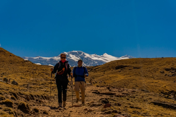Two hikers walking along a high-altitude mountain trail with snow-covered peaks in the background