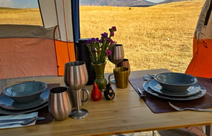 Table set for two inside a dining tent, with blue tableware, metal goblets and purple flowers.